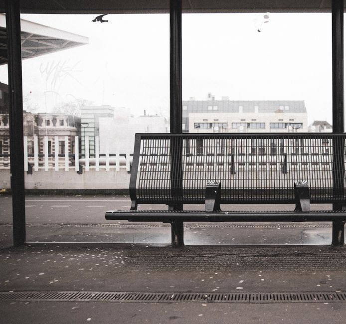 A black metal bench with a slatted seat and backrest positioned on an outdoor concrete platform or bus stop shelter. The bench is located in front of large glass panels that form part of a shelter, wi